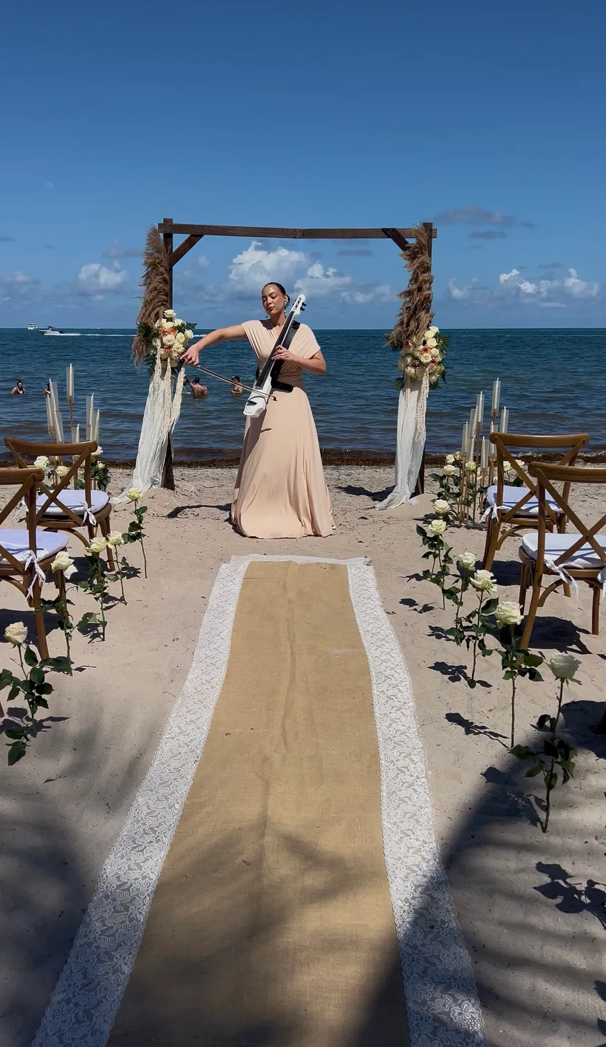 Saydee Sarduy playing electric cello beneath a floral arch at a beach ceremony.
